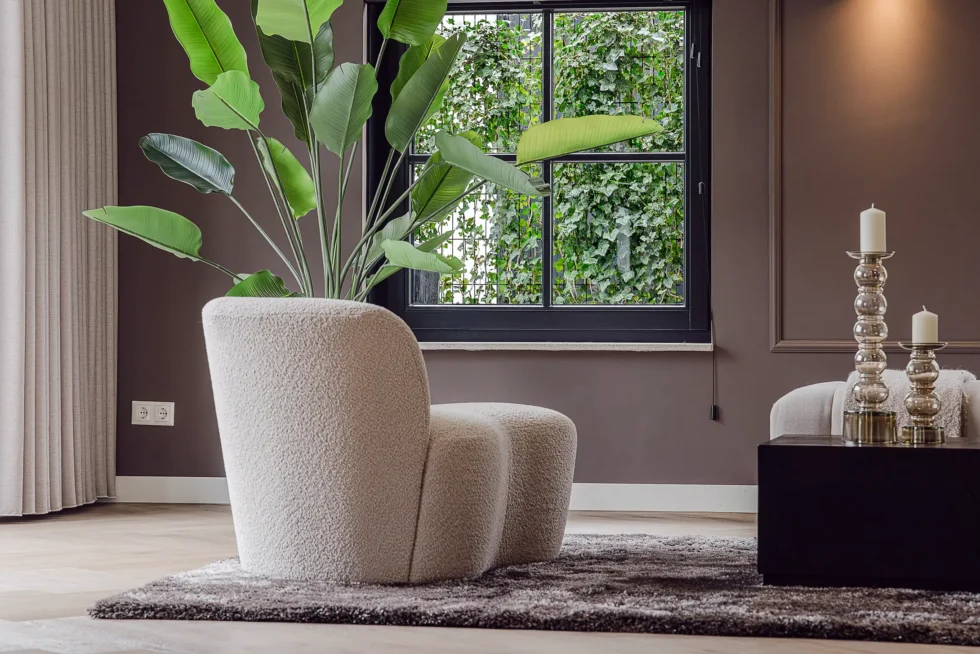 Zachte, beige bouclé fauteuil in de vorm van een vlinderstoel op een grijs vloerkleed, naast een grote bananenplant en een raam.