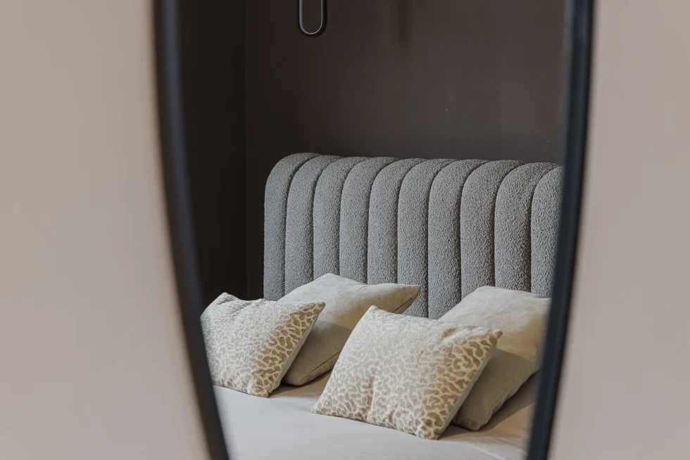 Reflectie in spiegel van een bed met gestoffeerd, verticaal geribbeld hoofdbord in grijs bouclé en gestapelde kussens.