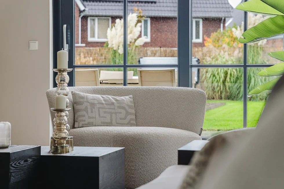 Laag bouclé fauteuil in crème met decoratief kussen, staand voor een groot glazen raam met uitzicht op de achtertuin.