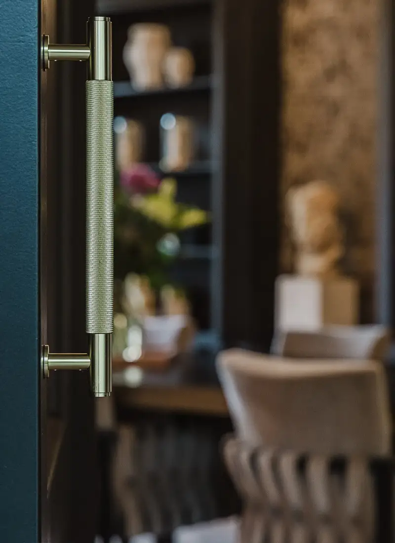 Close-up van een gedraaide deurklink op een blauwgroene deur, met een wazige doorkijk naar een eetkamer met beeld, bloemen en stoelen — Brikkr Exclusief