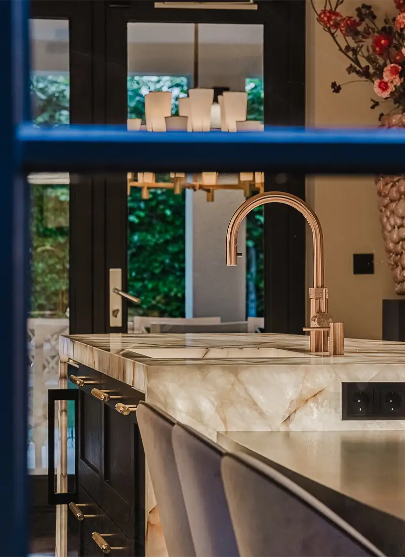 Sfeervolle interieurfoto van een luxe keuken met marmeren kookeiland, koperen kraan en zicht door openslaande deuren naar de tuin — Brikkr Exclusief